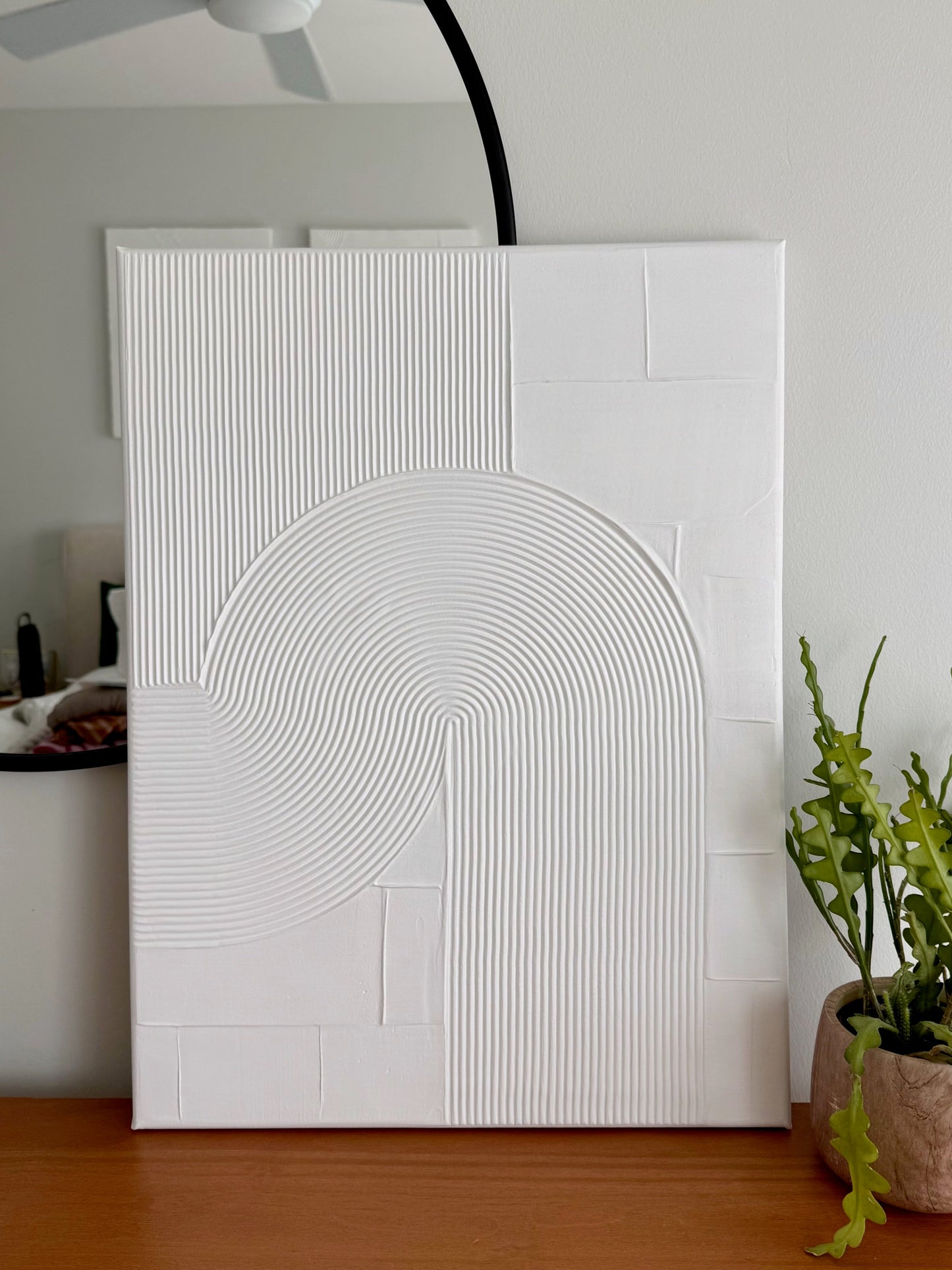 White textured wall art piece on a wooden floor with a plant and ceiling fan in the background.