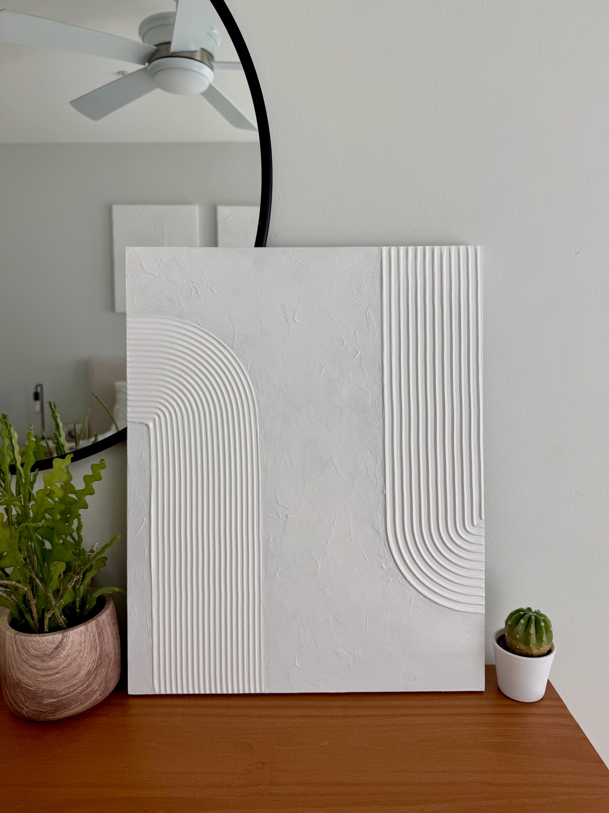 White textured wall art piece on a wooden surface with plants in the background