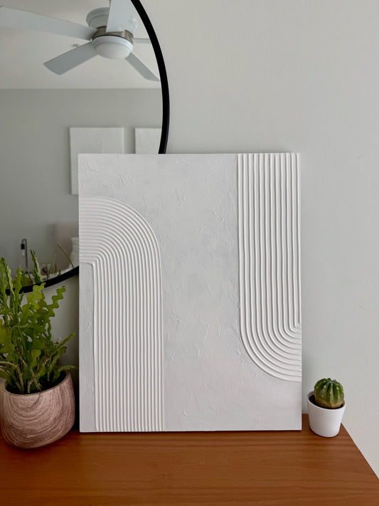 White textured wall art piece on a wooden surface with plants in the background