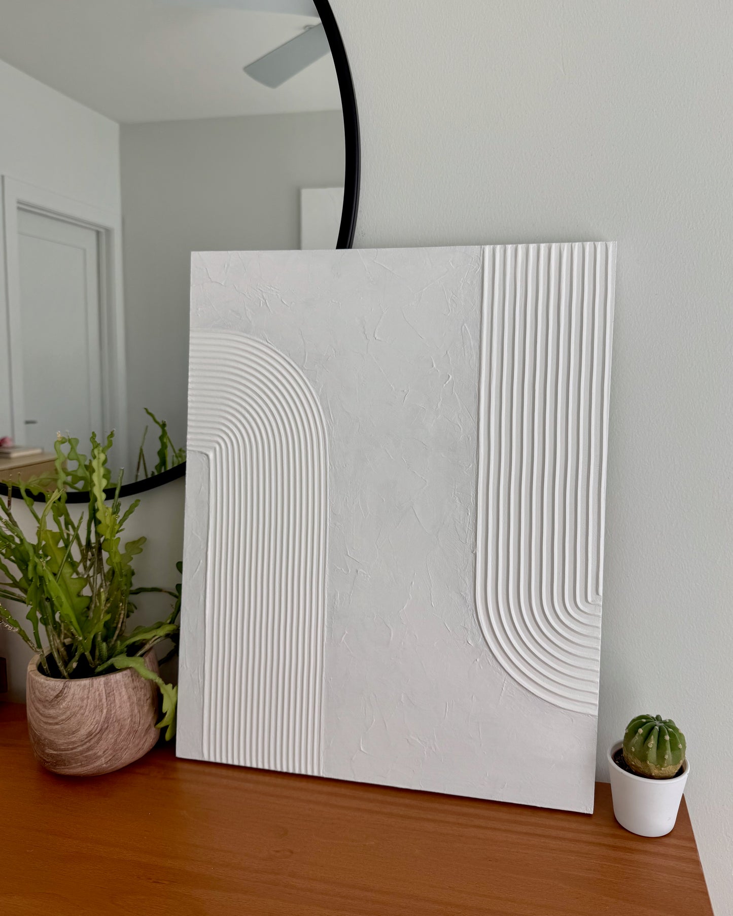 White textured wall art piece on a wooden surface with plants in the background