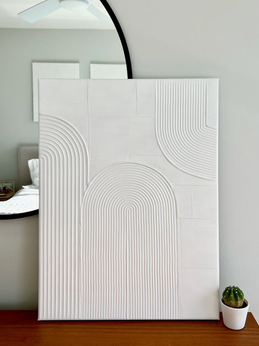 White textured wall art piece on a wooden surface with a mirror and plant in the background.