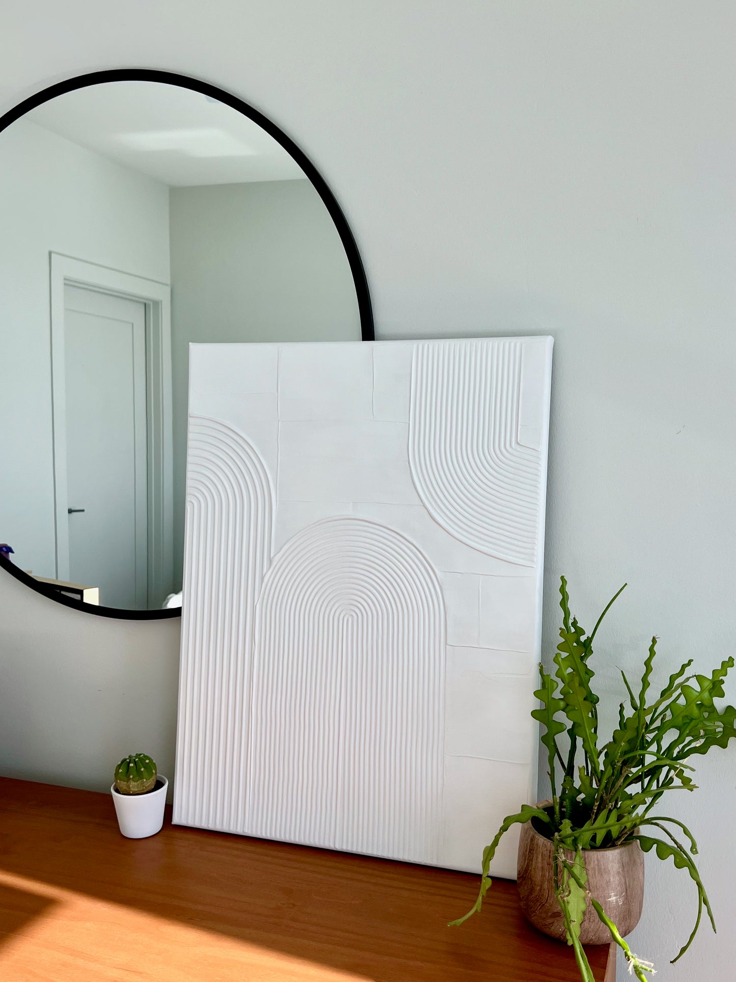 Round mirror on a wall with a white textured painting and potted plants below.