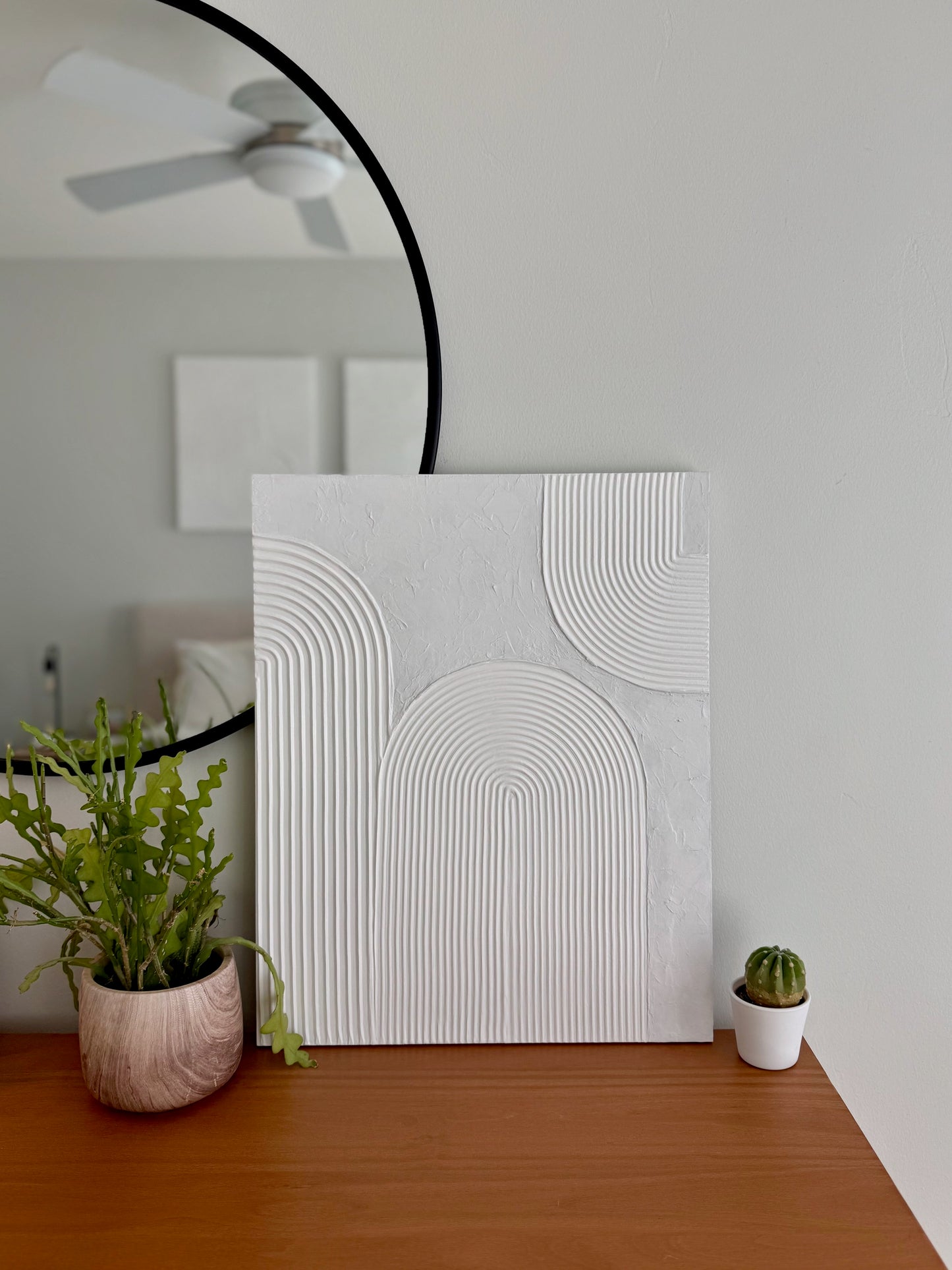 Abstract wall art piece on a wooden surface with plants and a mirror in the background.