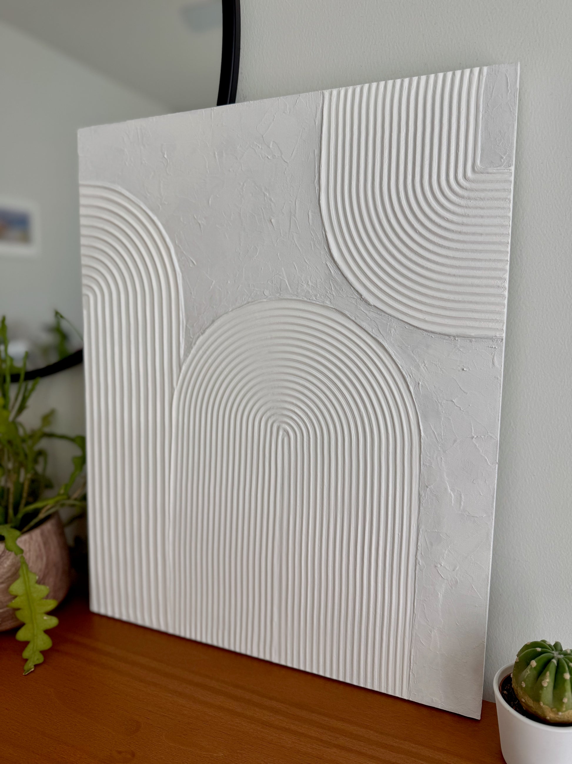 Textured white wall art piece on a wooden surface with plants in the background