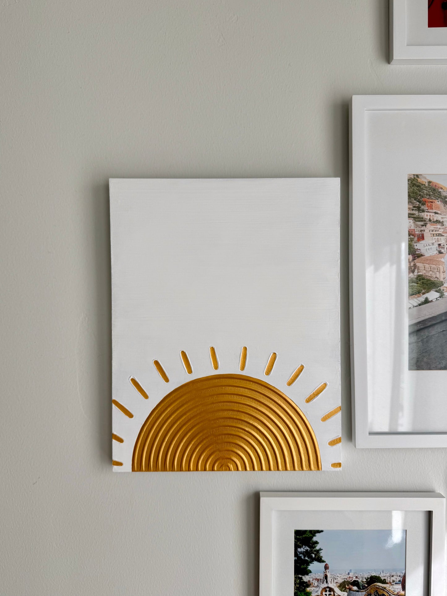 White wall with a gold sun design on a white canvas, surrounded by other framed pictures.