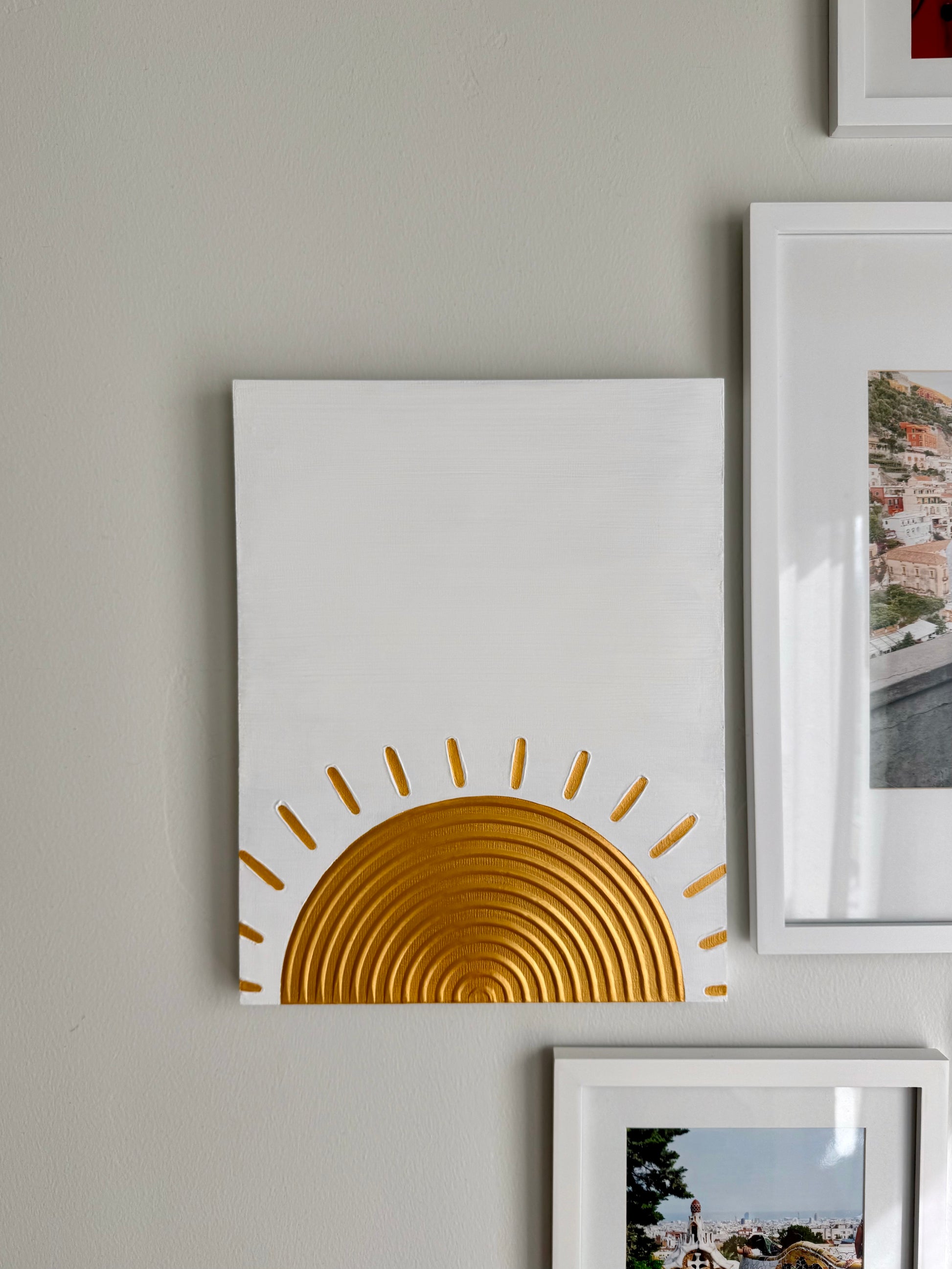 White wall with a gold sun design on a white canvas, surrounded by other framed pictures.