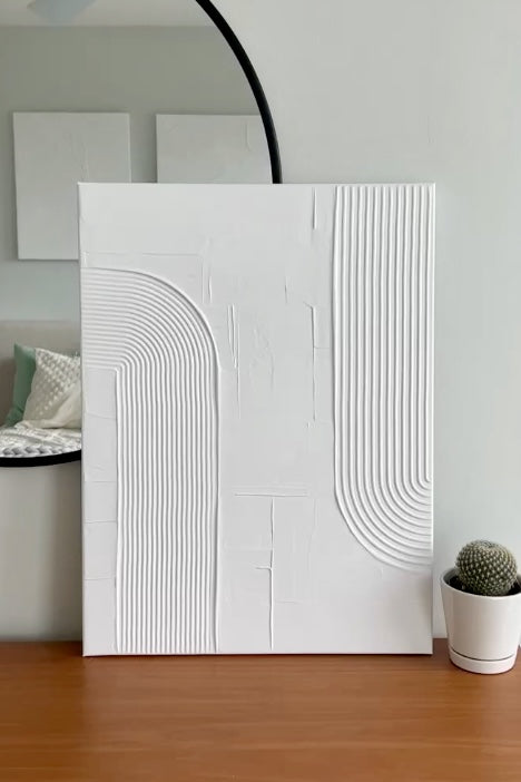 White textured wall art piece on a wooden surface with a plant in the background