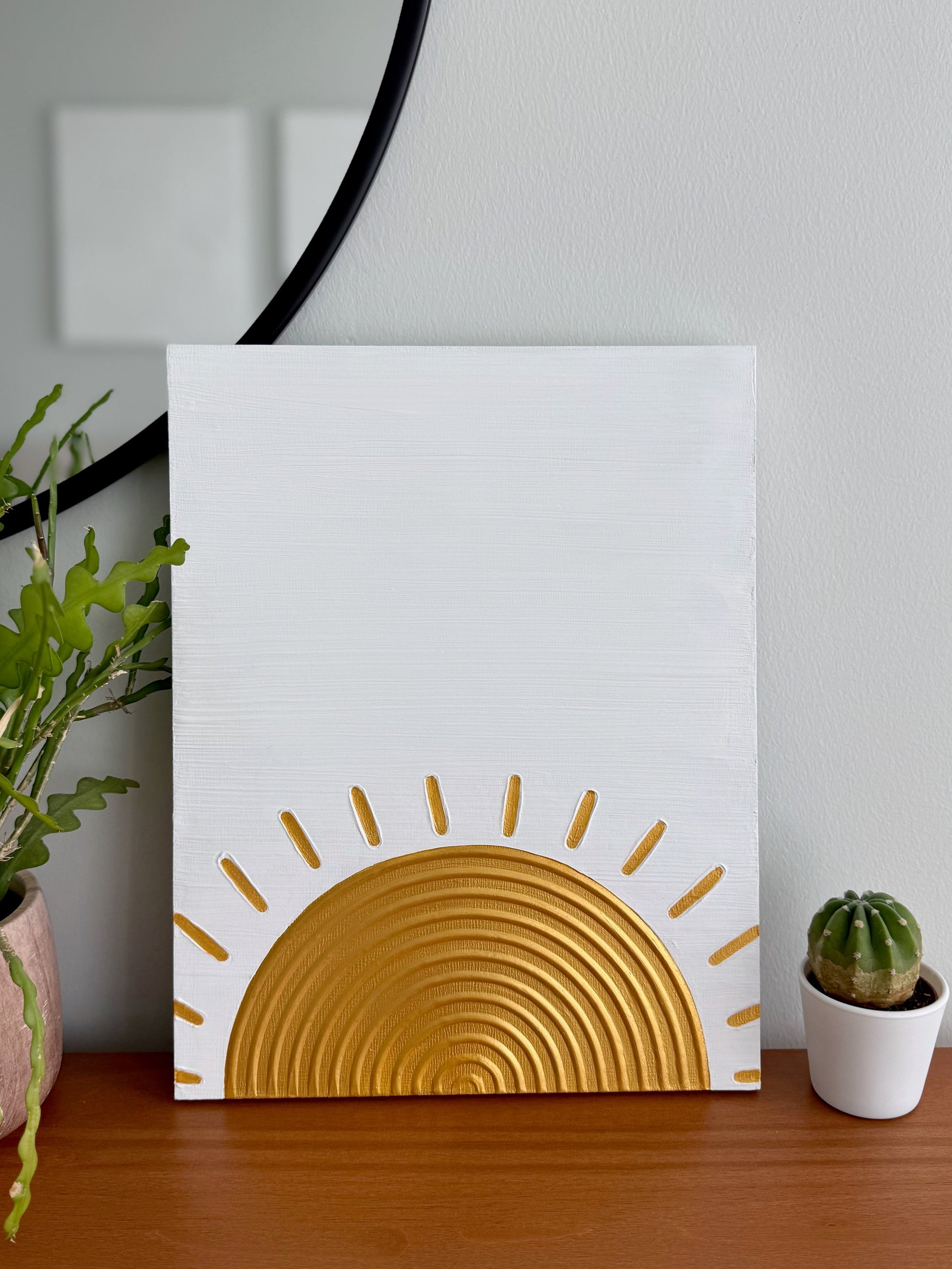 Decorative wall art with a gold sun design on a white background, placed on a wooden surface with plants.