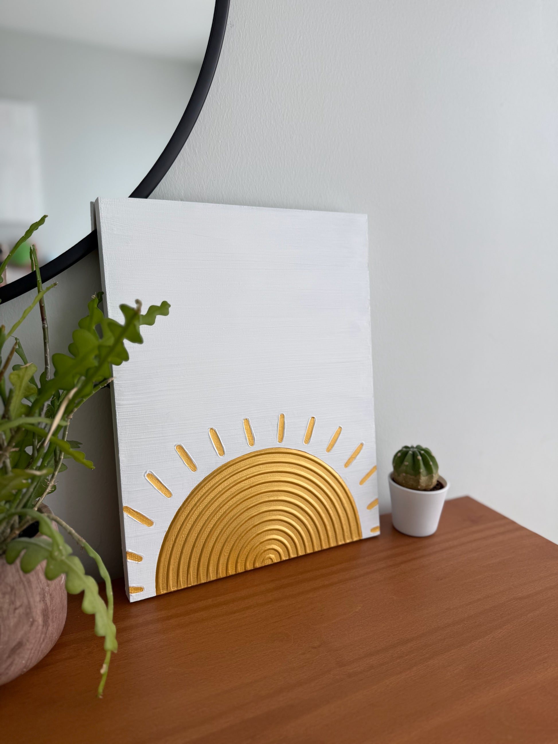 Decorative wall art with a gold sun design on a white background, placed on a wooden surface with plants.