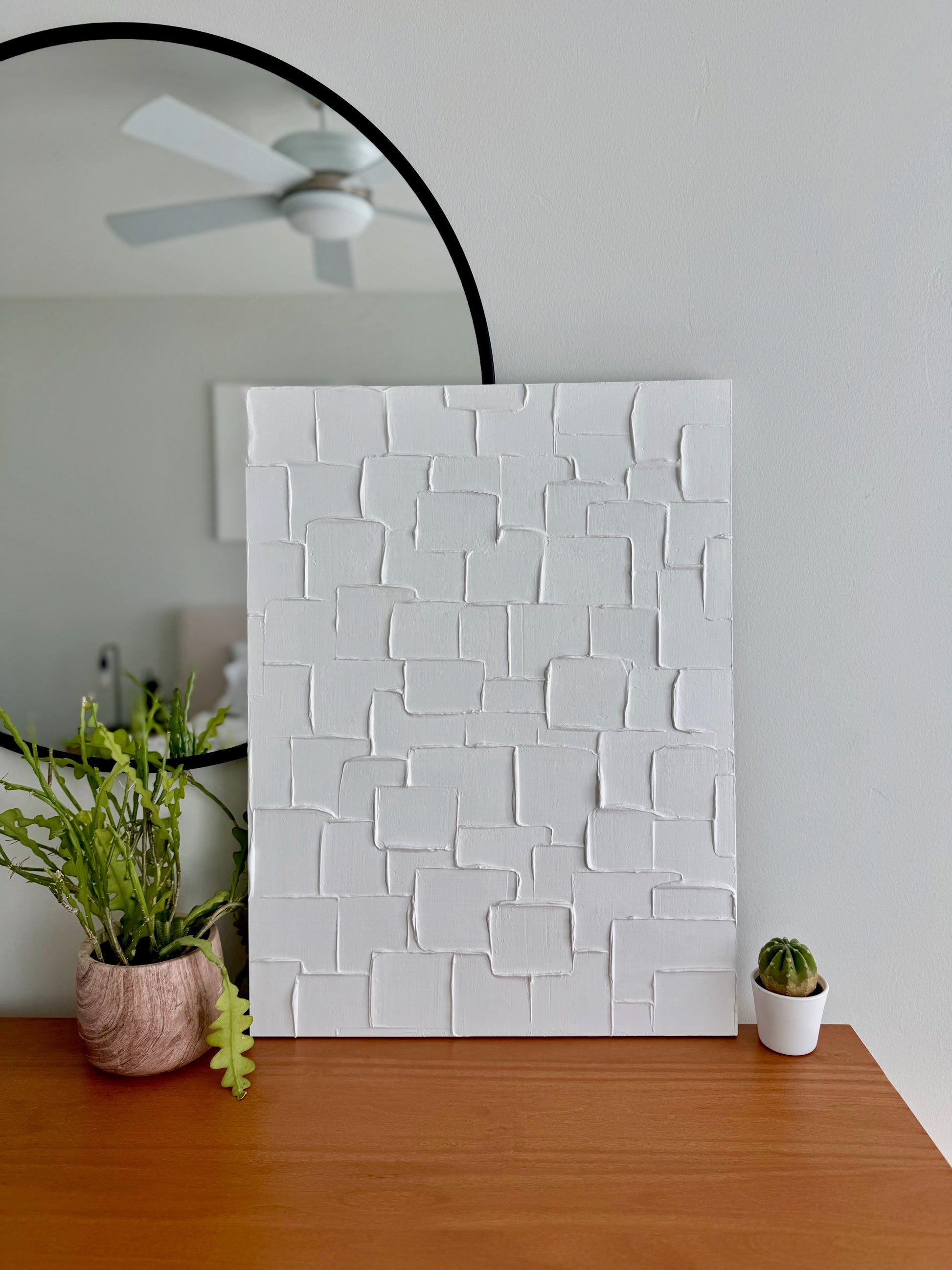 White textured wall panel on a wooden floor with plants and a mirror in the background.