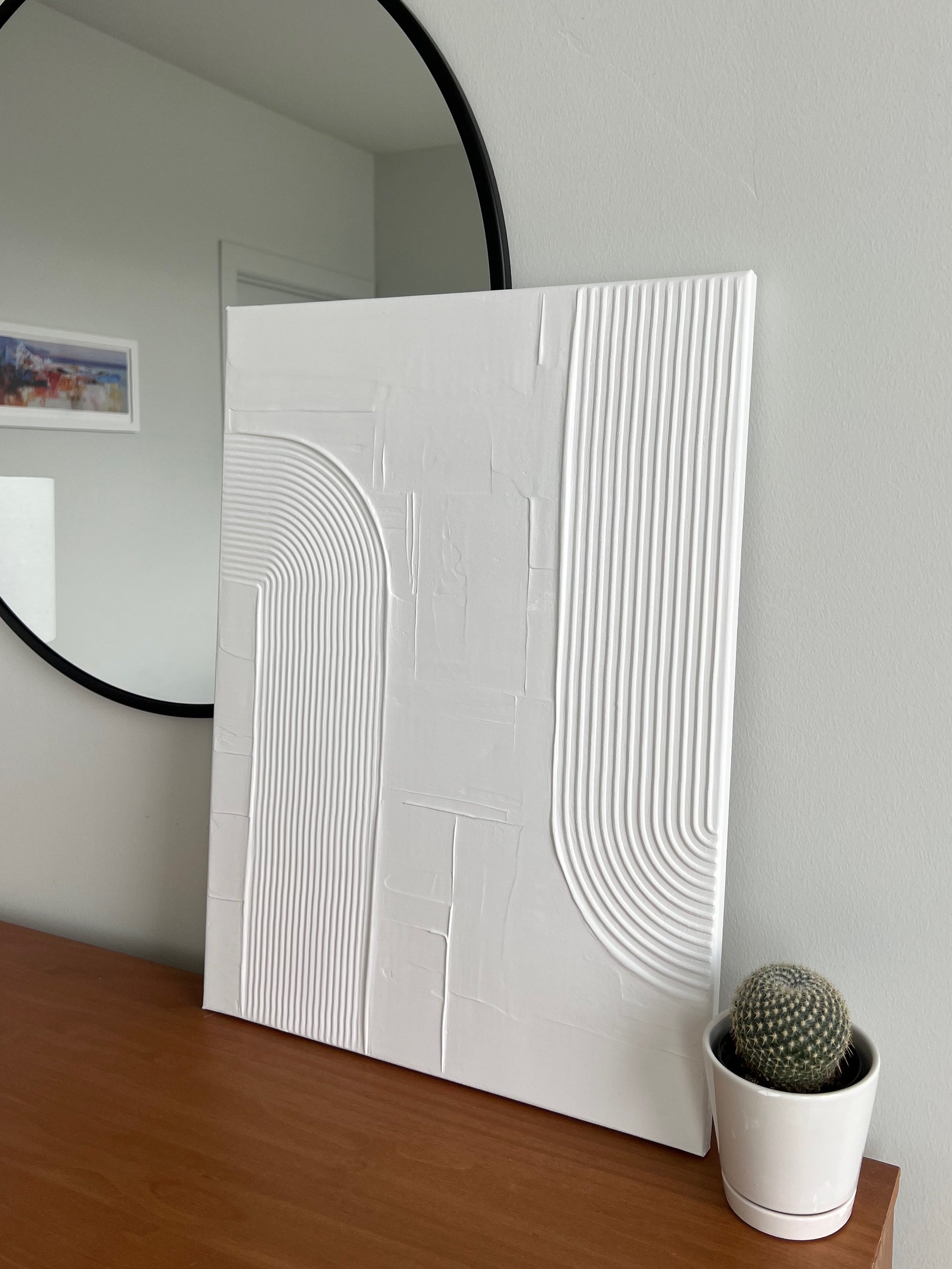 White textured wall art on a wooden shelf with a round mirror and small cactus in the background.