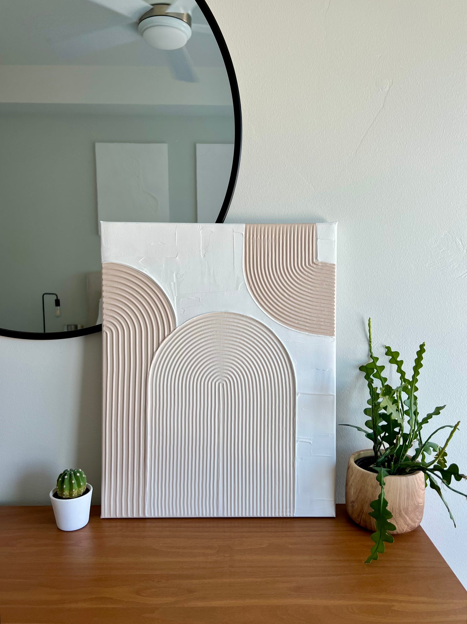 Abstract textured wall art on a wooden dresser with plants and a mirror in the background.