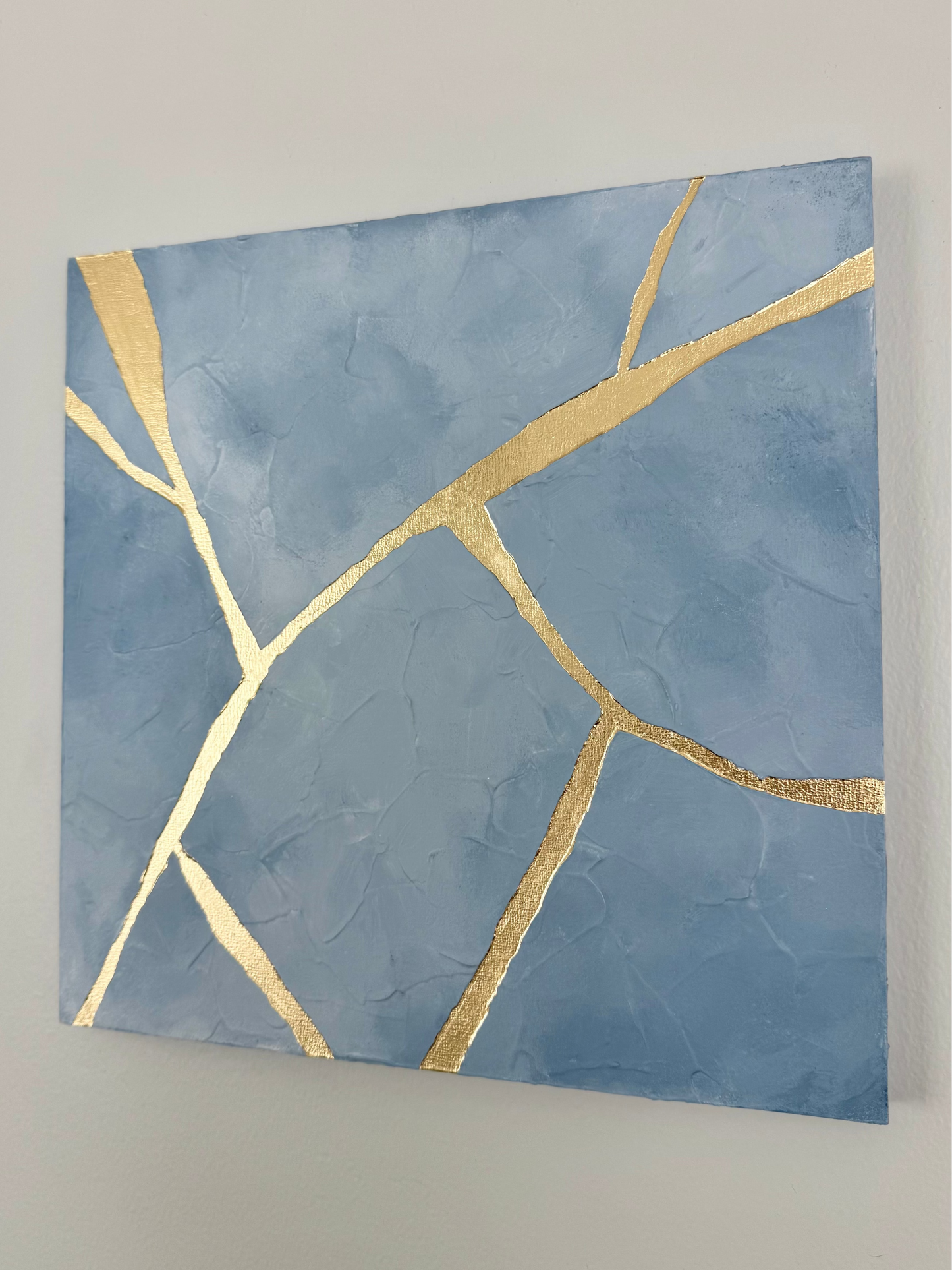 Square canvas wall art featuring a textured light blue background with metallic gold crackle lines, displayed on a neutral off-white wall.