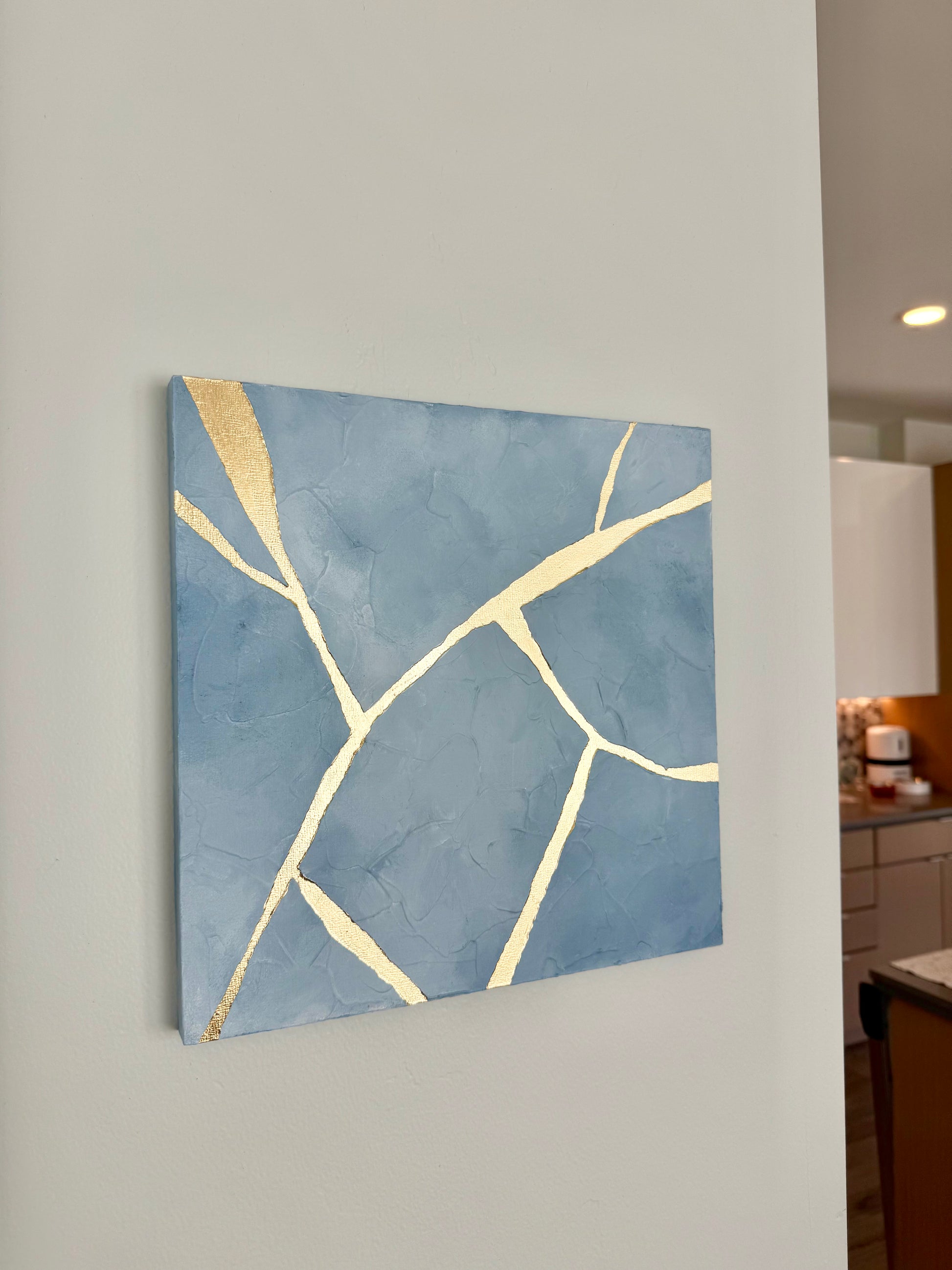 Square abstract wall art featuring a textured slate blue background with metallic gold leaf crackle lines, displayed on a light gray wall in a kitchen setting.
